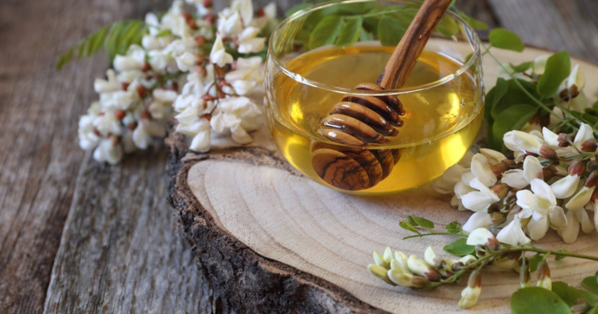 Pot de miel d’acacia très clair et presque translucide, ouvert sur un plan de travail clair, le miel s’écoulant en filet continu depuis une cuillère