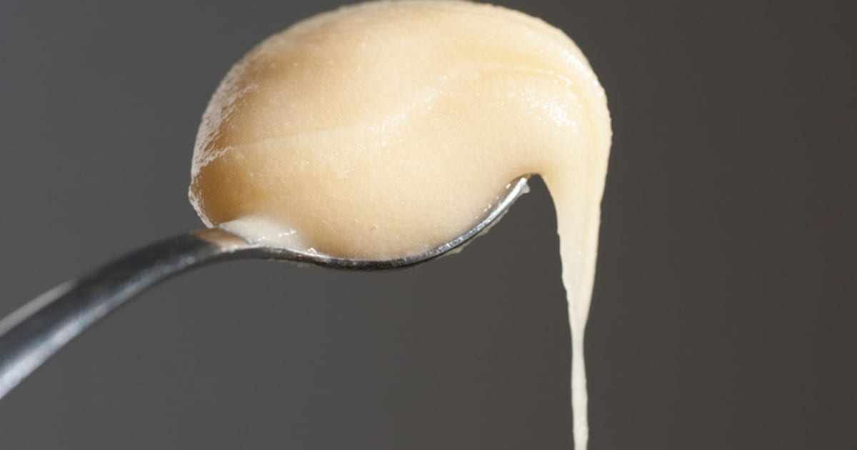 Pot ouvert de miel blanc crémeux à la texture opaque et satinée, cuillère en bois posée à côté sur un plan de travail clair